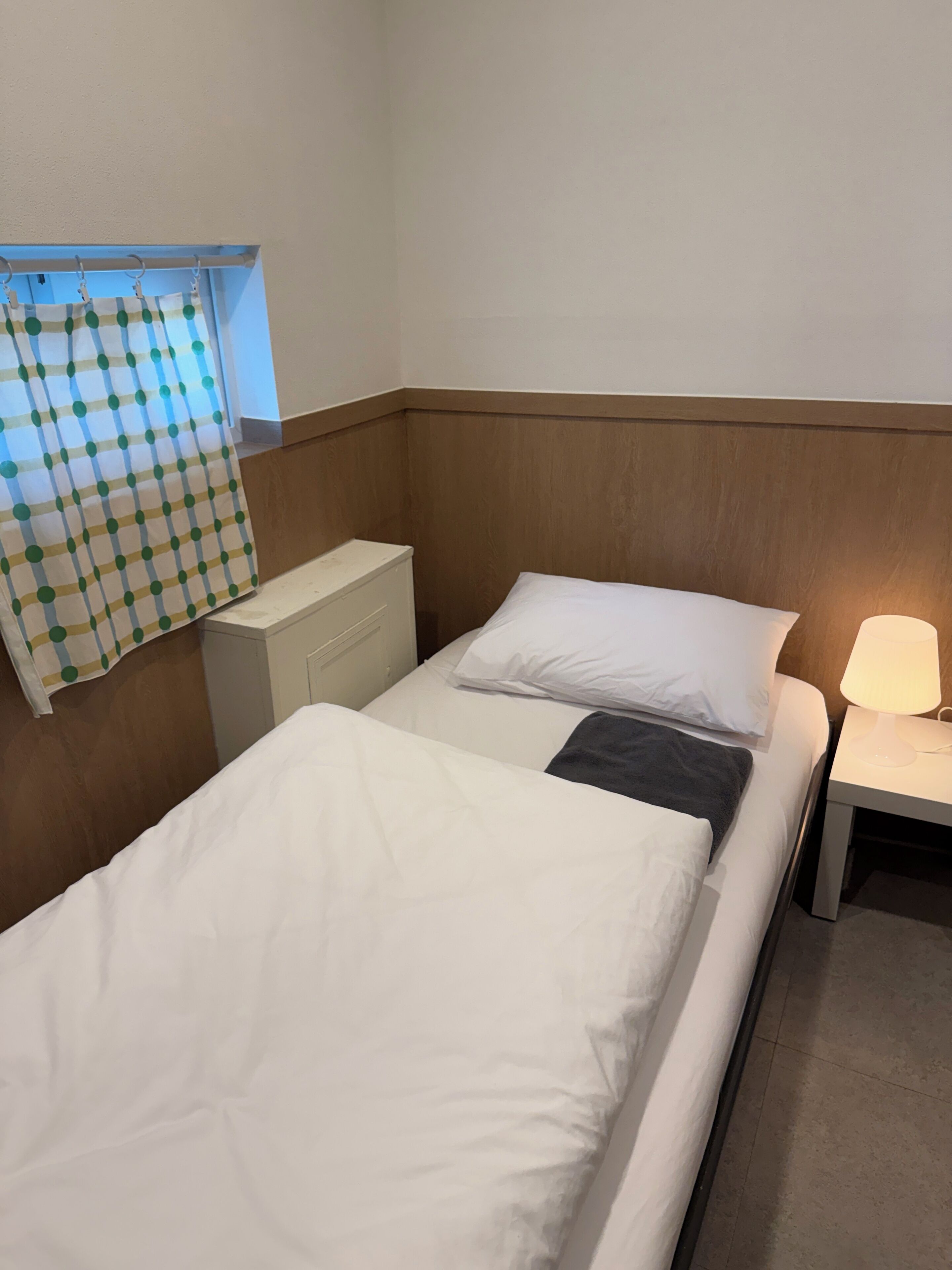 Een hotelkamer met twee eenpersoonsbedden, een nachtkastje met een lamp, een raam met een geruit gordijn en een houten hoofdbord.