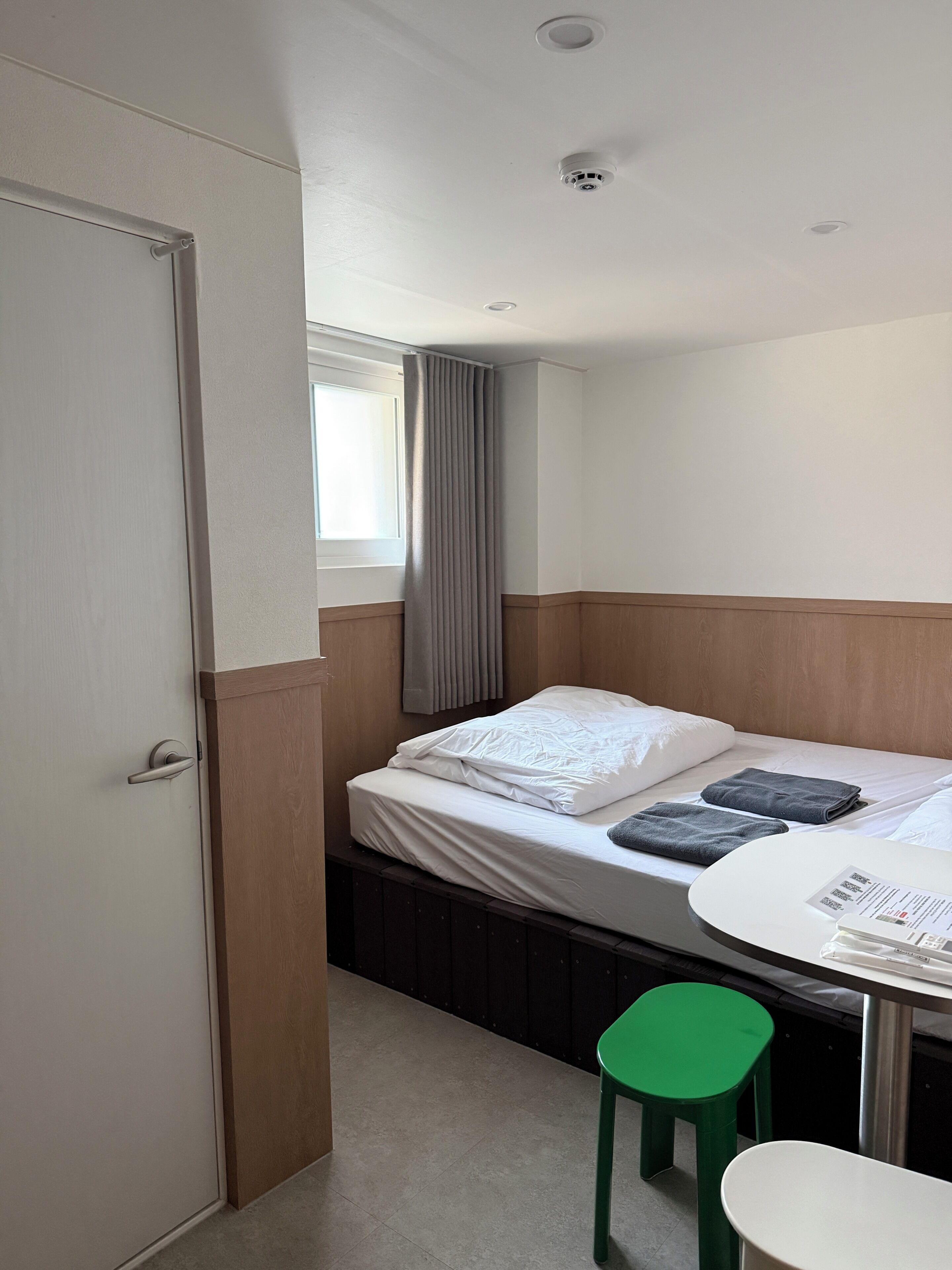 A hotel room with a bed, a table, a green stool, a window with curtains, and a door.