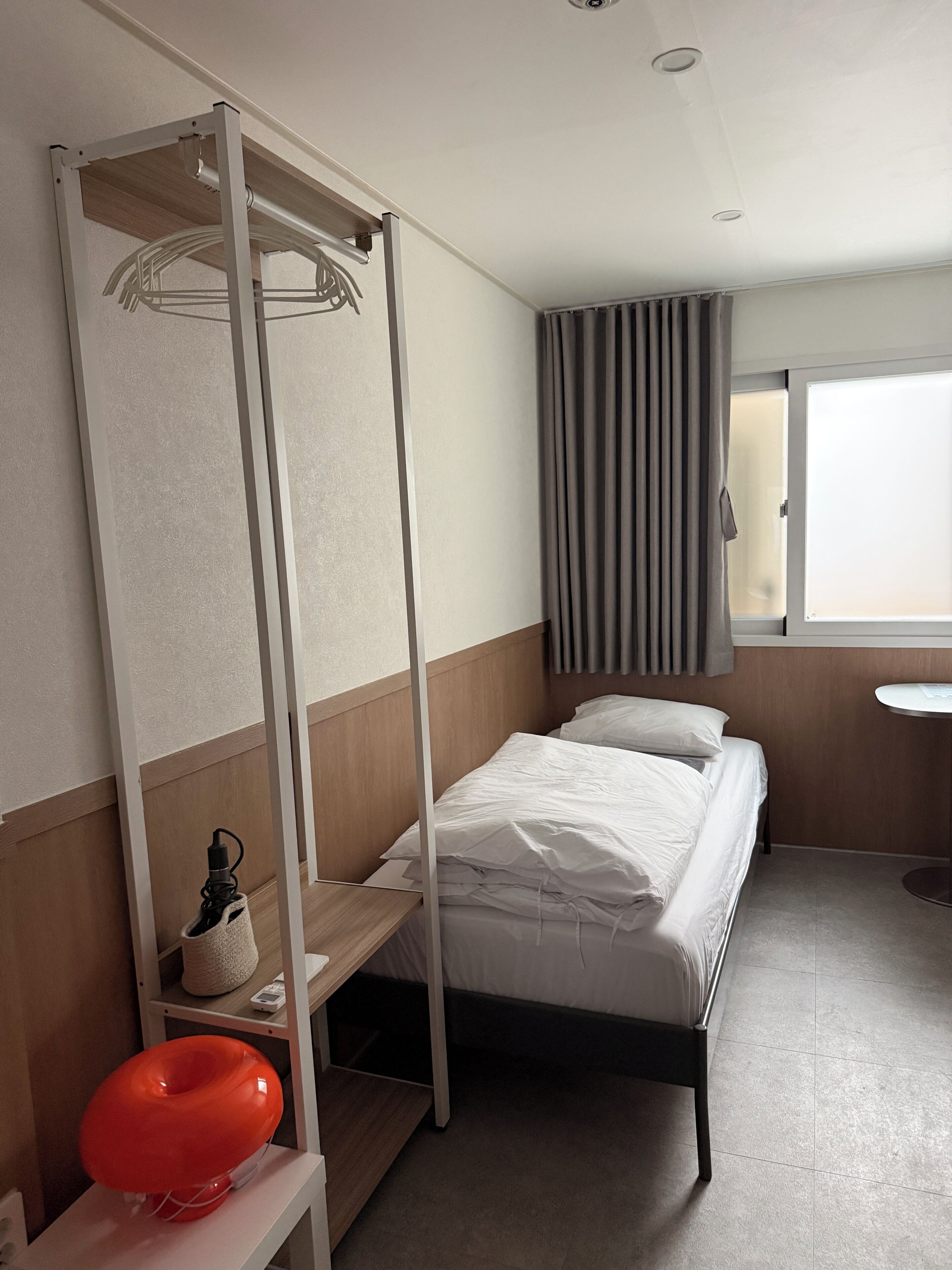 A hotel room with a bunk bed, a small table, a chair, a window with curtains, and a view of the outside.