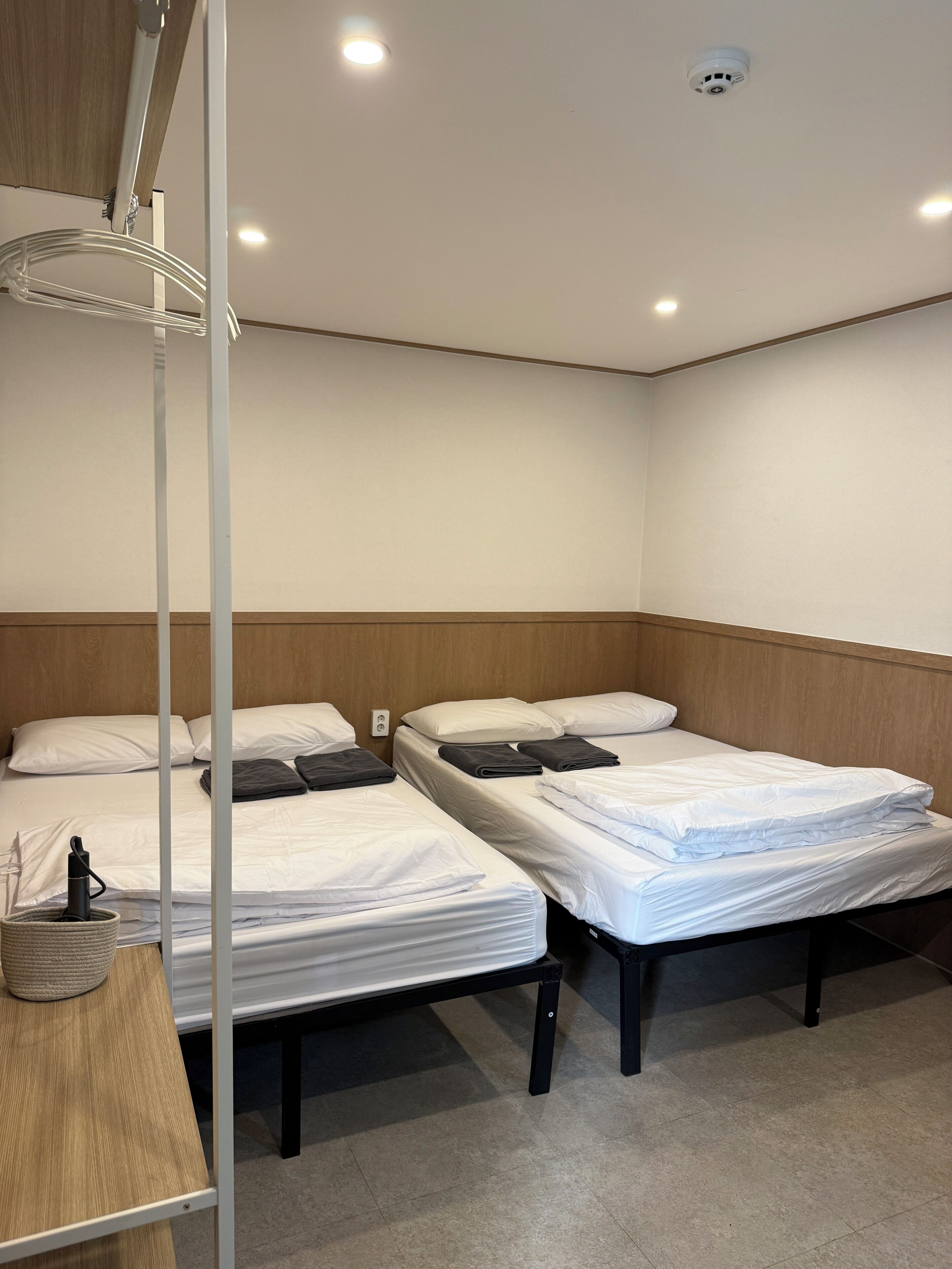 A hotel room with two beds, a bedside table, a lamp, and a ceiling-mounted security camera.