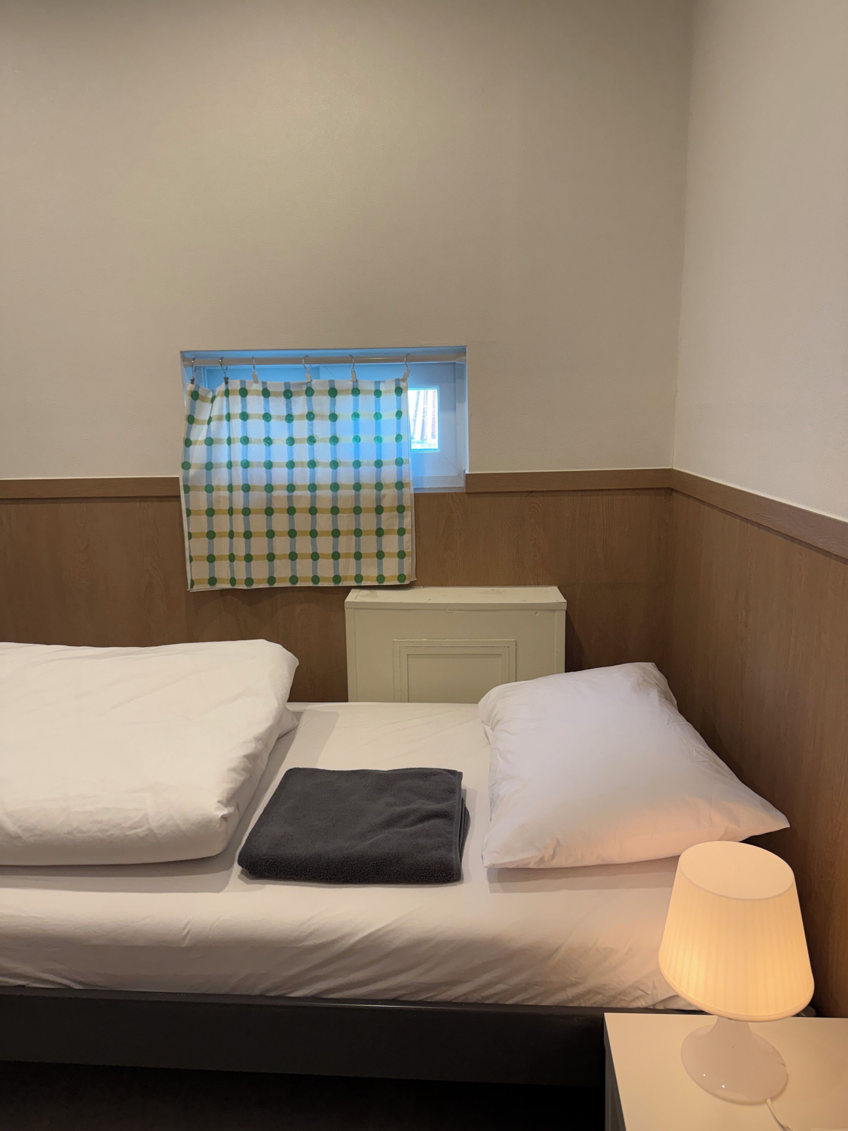 A single bed with white linens, a black towel, a window with a checkered curtain, and a bedside lamp.