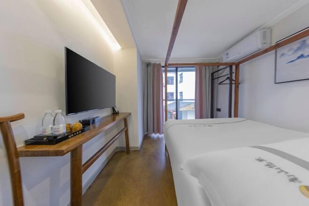 Quarto de hotel com cama, televisão de tela plana, escrivaninha de madeira com bandeja de objetos e janela com vista para edifícios.