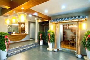 A hotel reception area with a wooden reception desk, potted plants, and a seating area with tables and chairs.