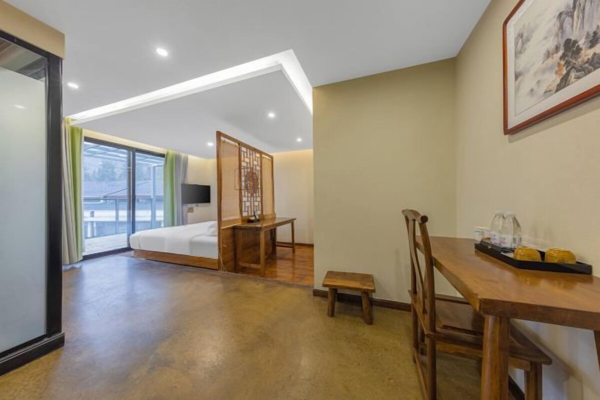 A hotel room with a bed, a desk, a small table, and a chair. There is a window with curtains and a view of the outside.