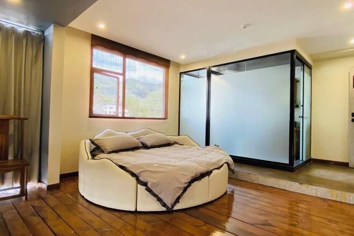 Uma cama redonda com cobertura bege em um quarto com piso de madeira, uma janela com vista e uma porta de correr de vidro.