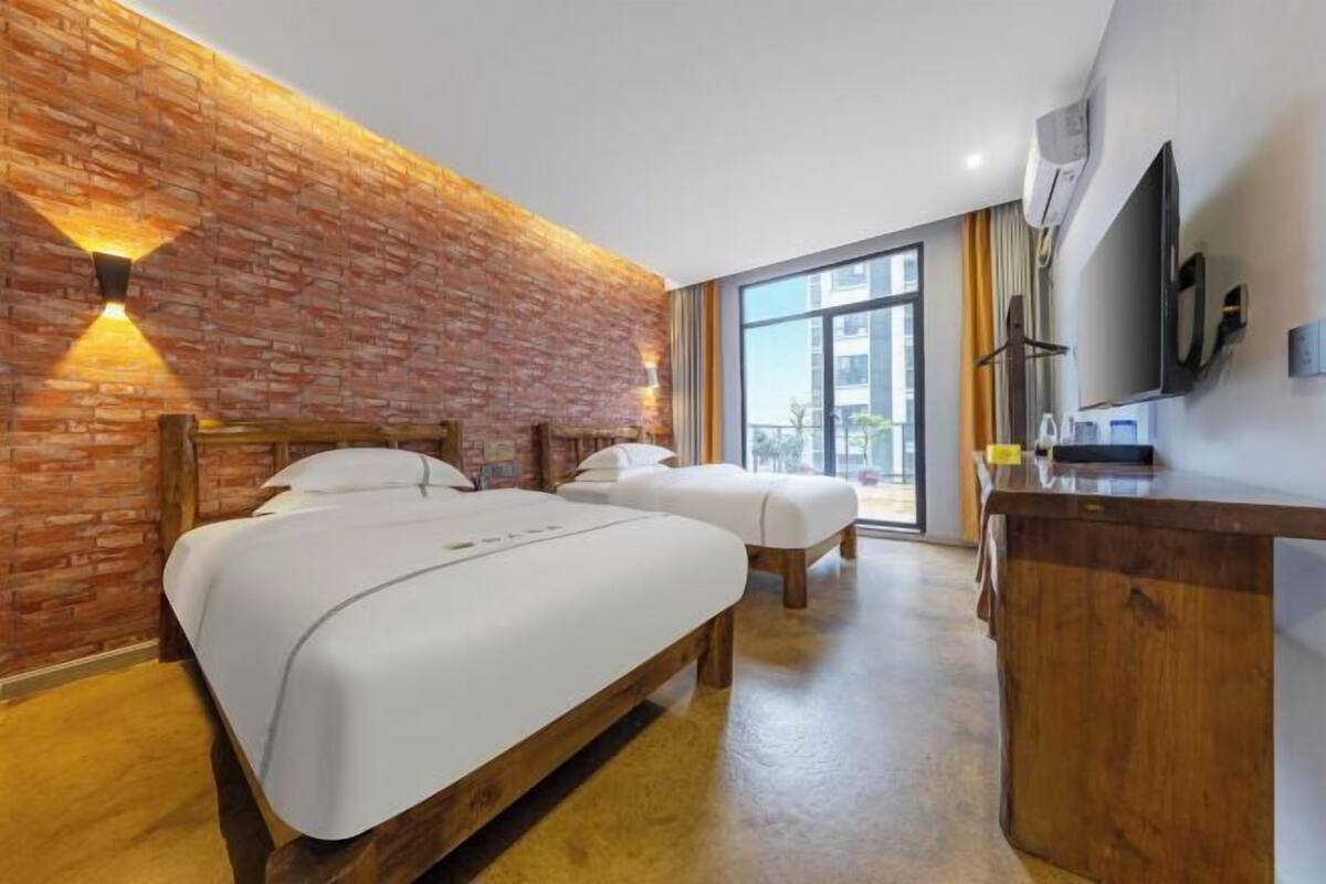 Quarto de hotel com duas camas, uma escrivaninha de madeira, televisão e parede de tijolos.