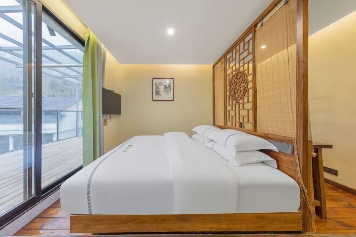 Quarto de hotel com cama, televisão, divisória de madeira e janela com vista para o exterior.