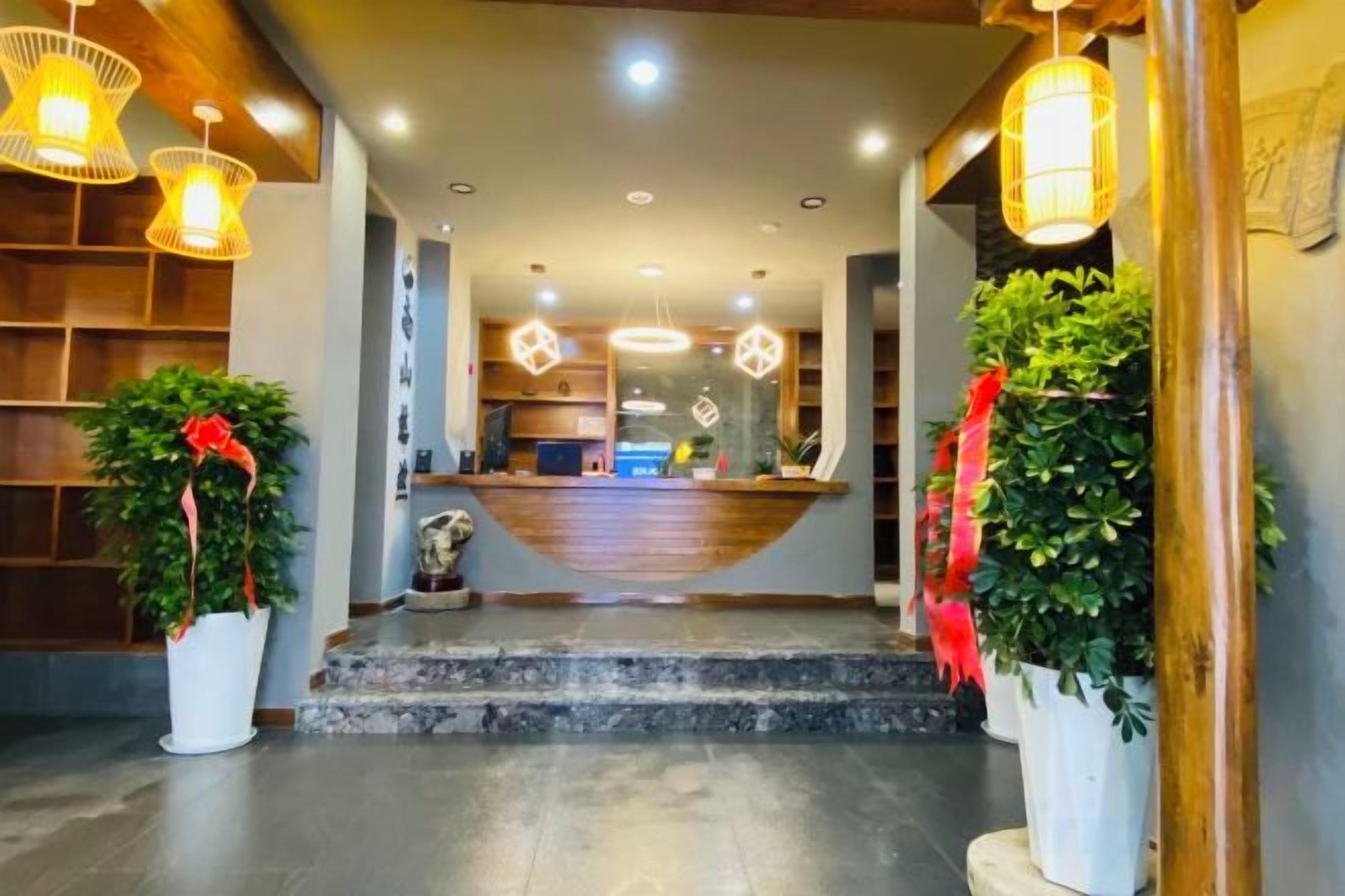 Um saguão de hotel com balcão de recepção, plantas em vasos e escada para o andar superior.