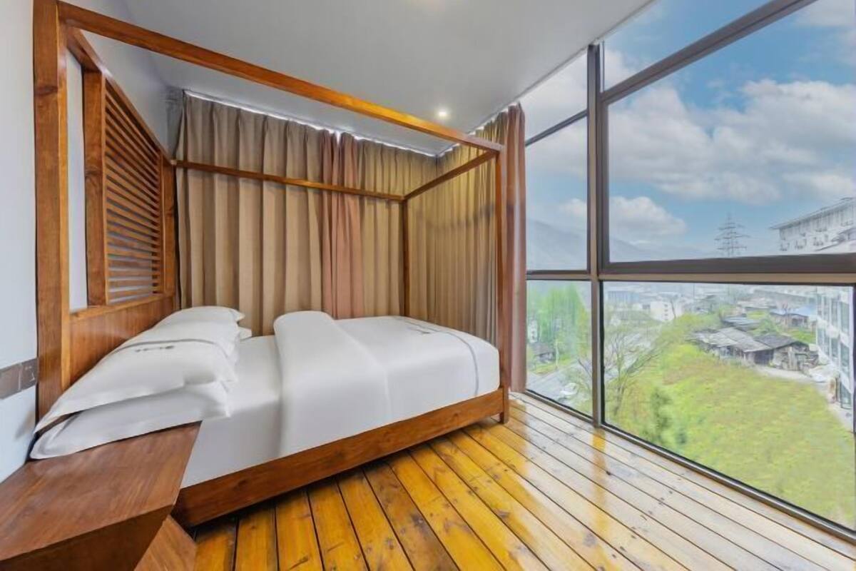 Quarto com estrutura de cama de madeira, vista para o horizonte urbano através de amplas janelas e teto inclinado.