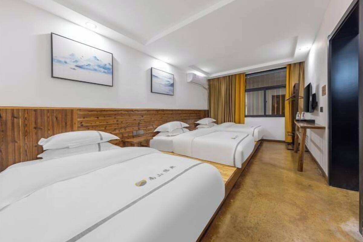 Quarto de hotel com três camas, cabeceiras de madeira, roupa de cama branca e uma janela grande com cortinas.
