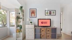 A modern living room with a television displaying "GUEST READY", a bookshelf, a plant, and a view of the outdoors. - GuestReady - Lovely haven with a private garden (London)