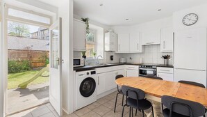 A modern kitchen with white cabinets, a wooden dining table, and a washing machine. - GuestReady - Lovely haven with a private garden (London)