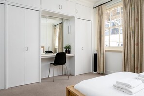 A bedroom with a bed, a desk with a chair, a wardrobe, and a window with curtains. - GuestReady - Convenient stay near Hyde Park (London)