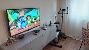 Ein Wohnzimmer mit Fernseher, der eine Zeichentricksendung zeigt, einem stationären Fahrrad und einem gemusterten Sessel.