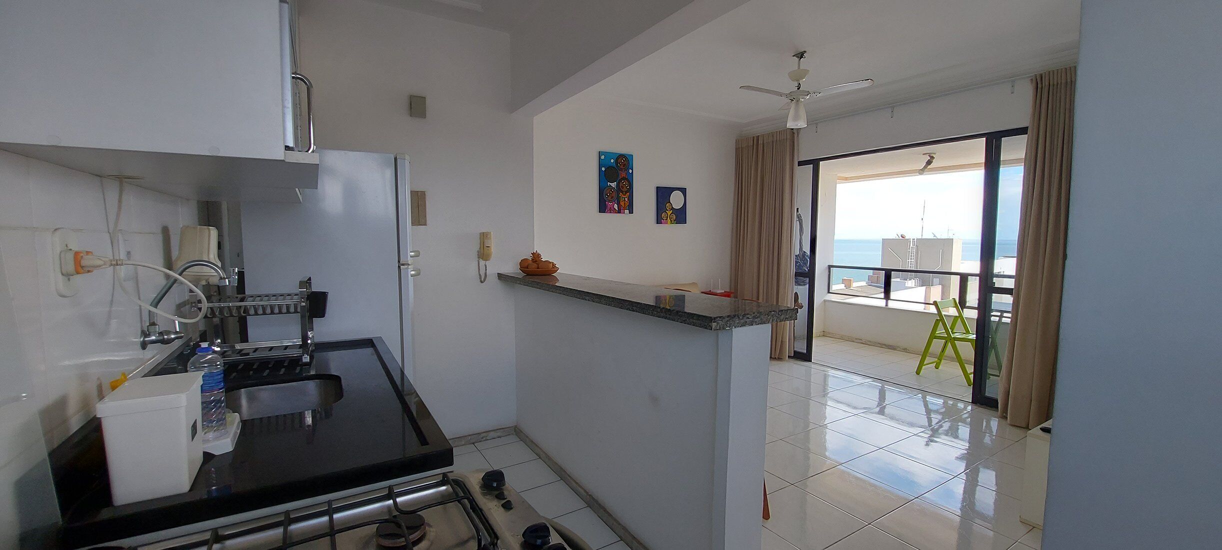 Cocina compacta con estufa a gas, un lavamanos y una pequeña encimera. Hay un balcón con vistas al mar y una silla verde.