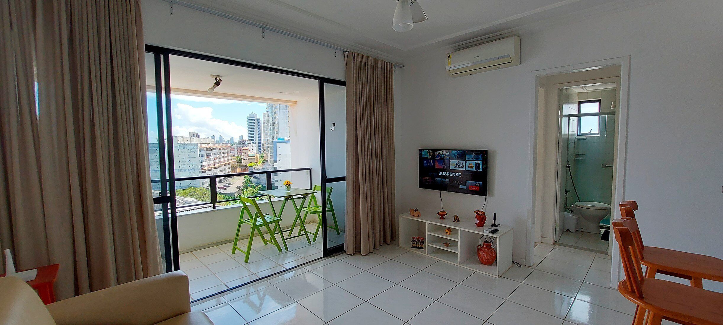 A modern living room with a large window, a flat-screen TV, and a balcony with green chairs.