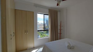 A bedroom with a bed, a wardrobe, a window with a view of buildings, and a coat rack. - CORVE0104 - Marítima Residence (Salvador)