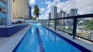 A rooftop pool with a city view, a palm tree, and a modern building with blue tiles. - CORVE0104 - Marítima Residence (Salvador)