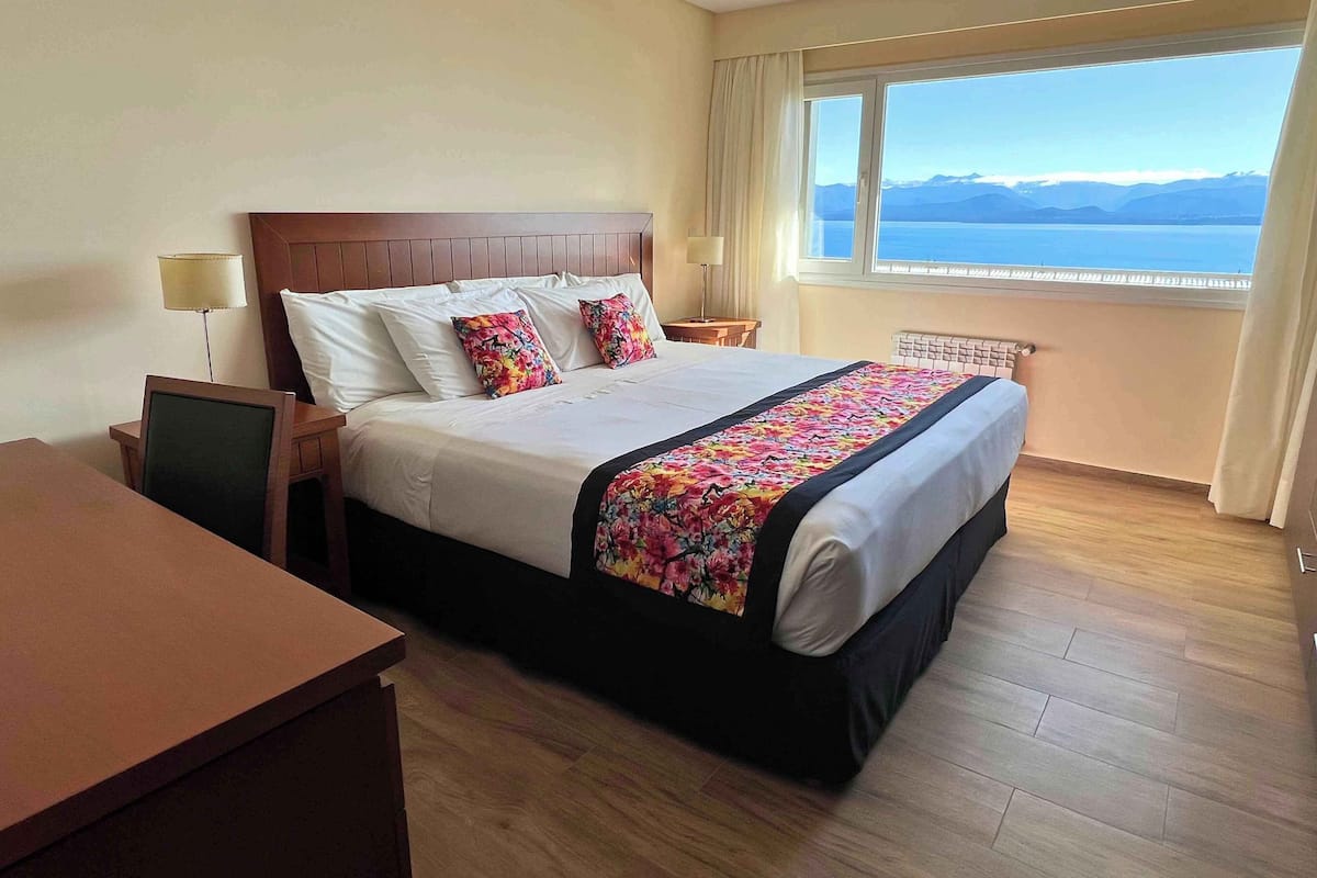 Une chambre d’hôtel avec un grand lit, un bureau, une chaise, une table de chevet, une lampe, une fenêtre donnant sur la mer et les montagnes, et un radiateur.