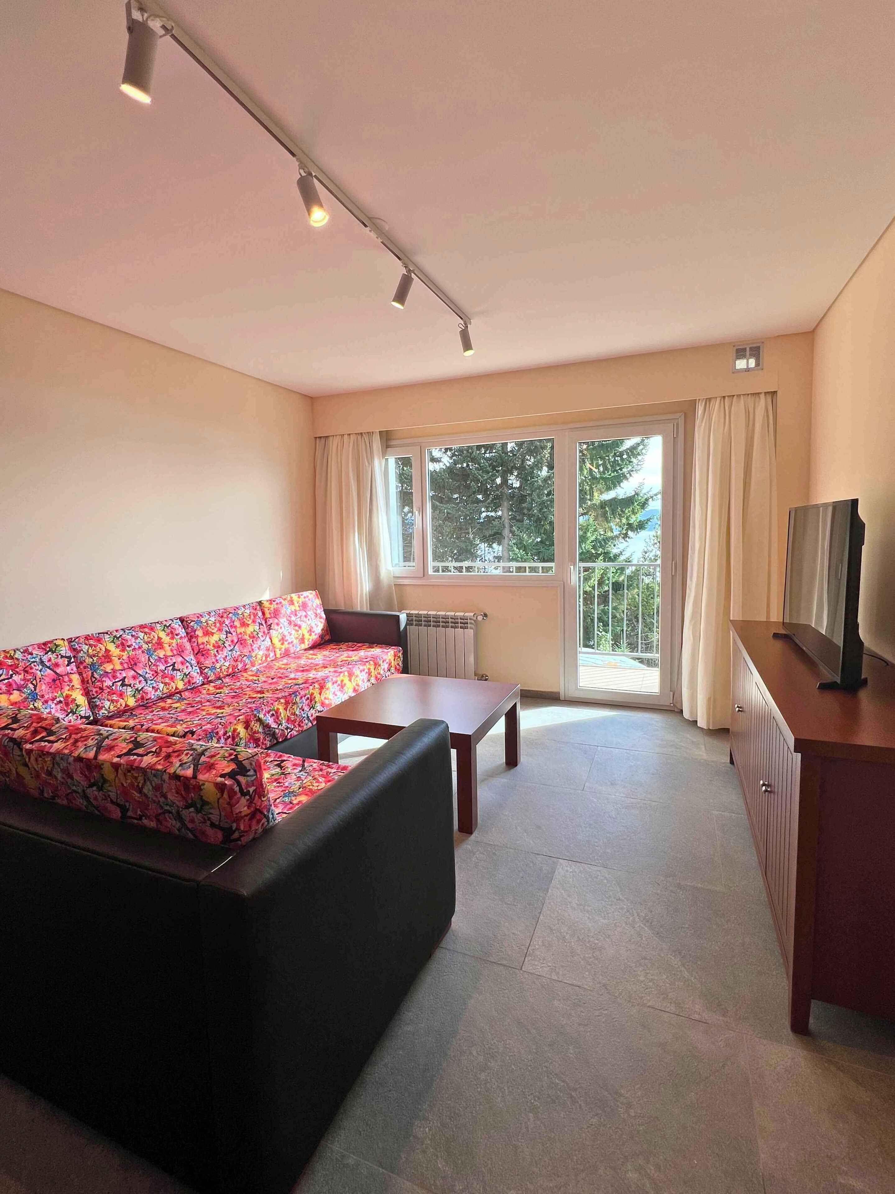 Ein Wohnzimmer mit einer Couch, einem Couchtisch, einem Fernseher auf einem Schrank und einem großen Fenster mit Vorhängen.