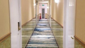 Een hotelgang met een kamerbreed tapijt, witte muren en deuren met een gouden embleem.