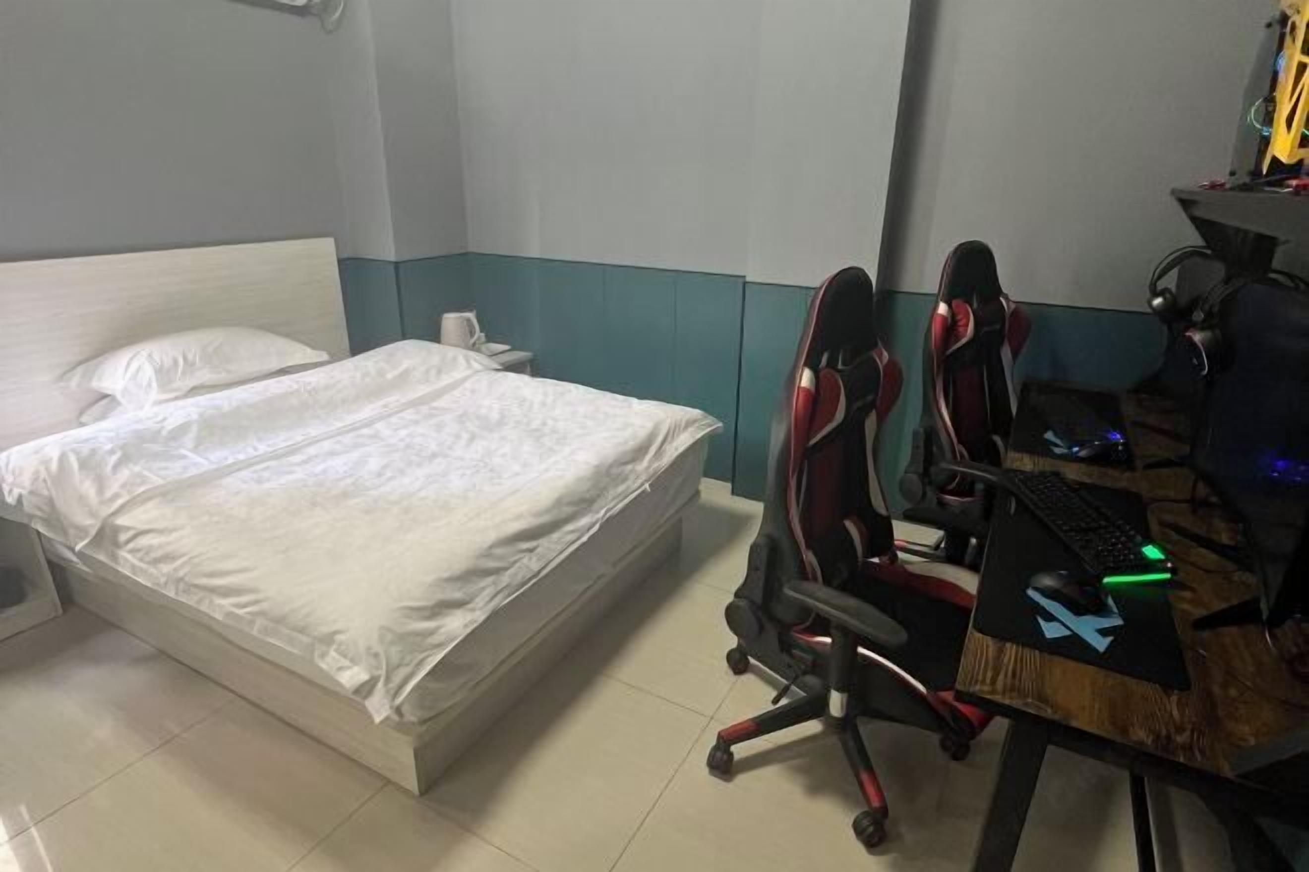 Habitación de hotel con una cama, un escritorio con computadora y silla gamer, y una pequeña mesa con teclado y ratón.