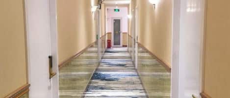 A hotel corridor with a carpeted floor, doors on either side, and a light fixture on the wall.