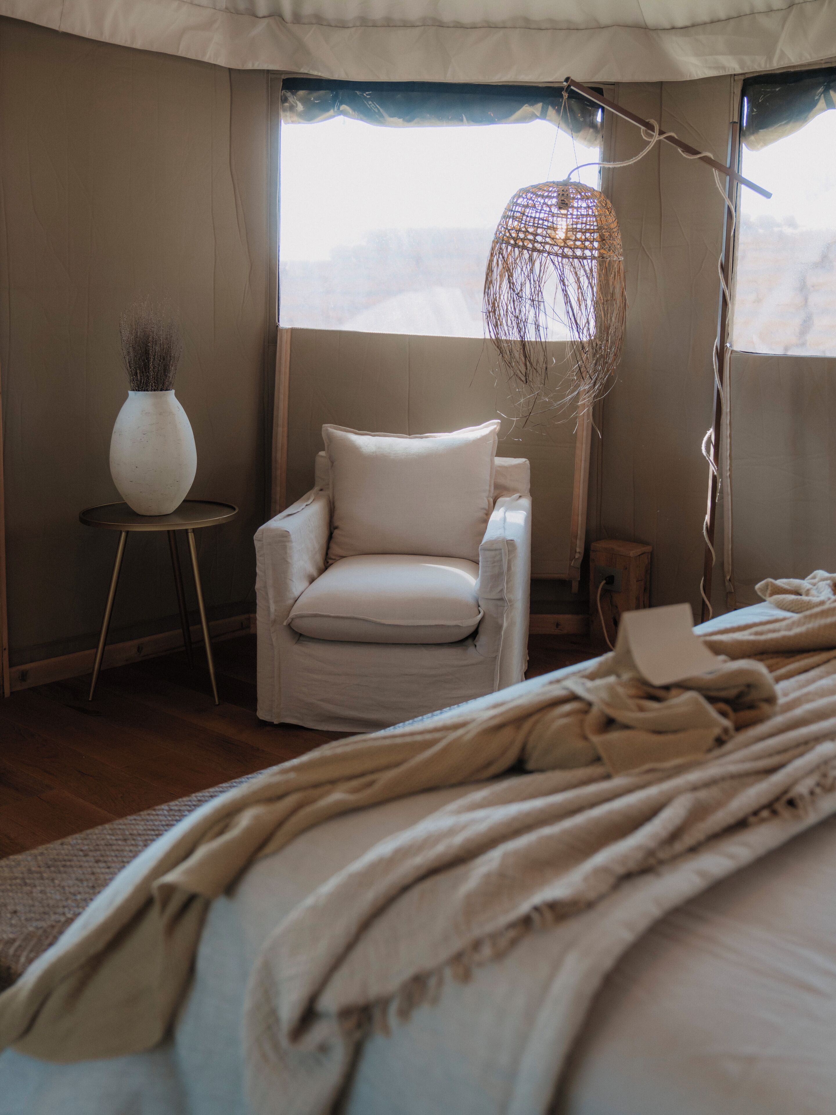 A bedroom with a bed, a white armchair, a small round table with a vase, and a hanging light fixture.