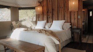 A large bed with white linens, a wooden headboard, and a bench in front. There are two wall-mounted lamps and a window with a view of trees. - UNAI Atacama Luxury Tents (San Pedro de Atacama)
