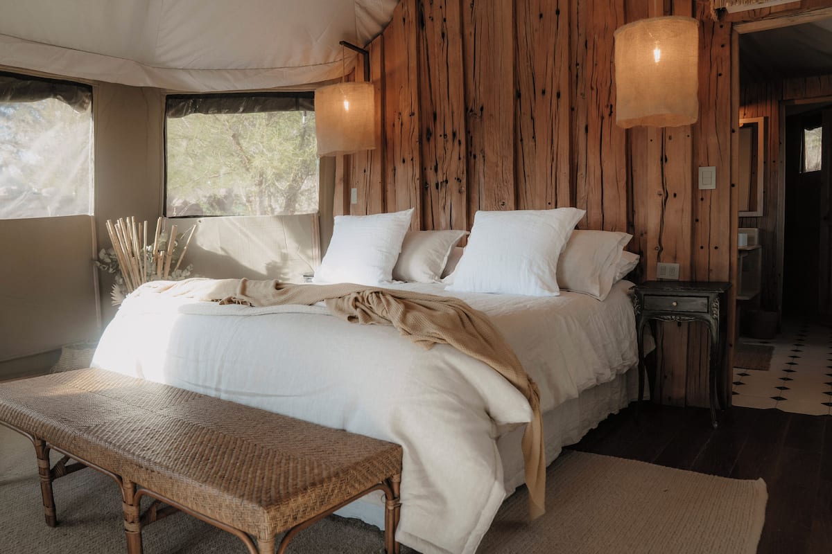 A large bed with white linens, a wooden headboard, and a bench in front. There are two wall-mounted lamps and a window with a view of trees.