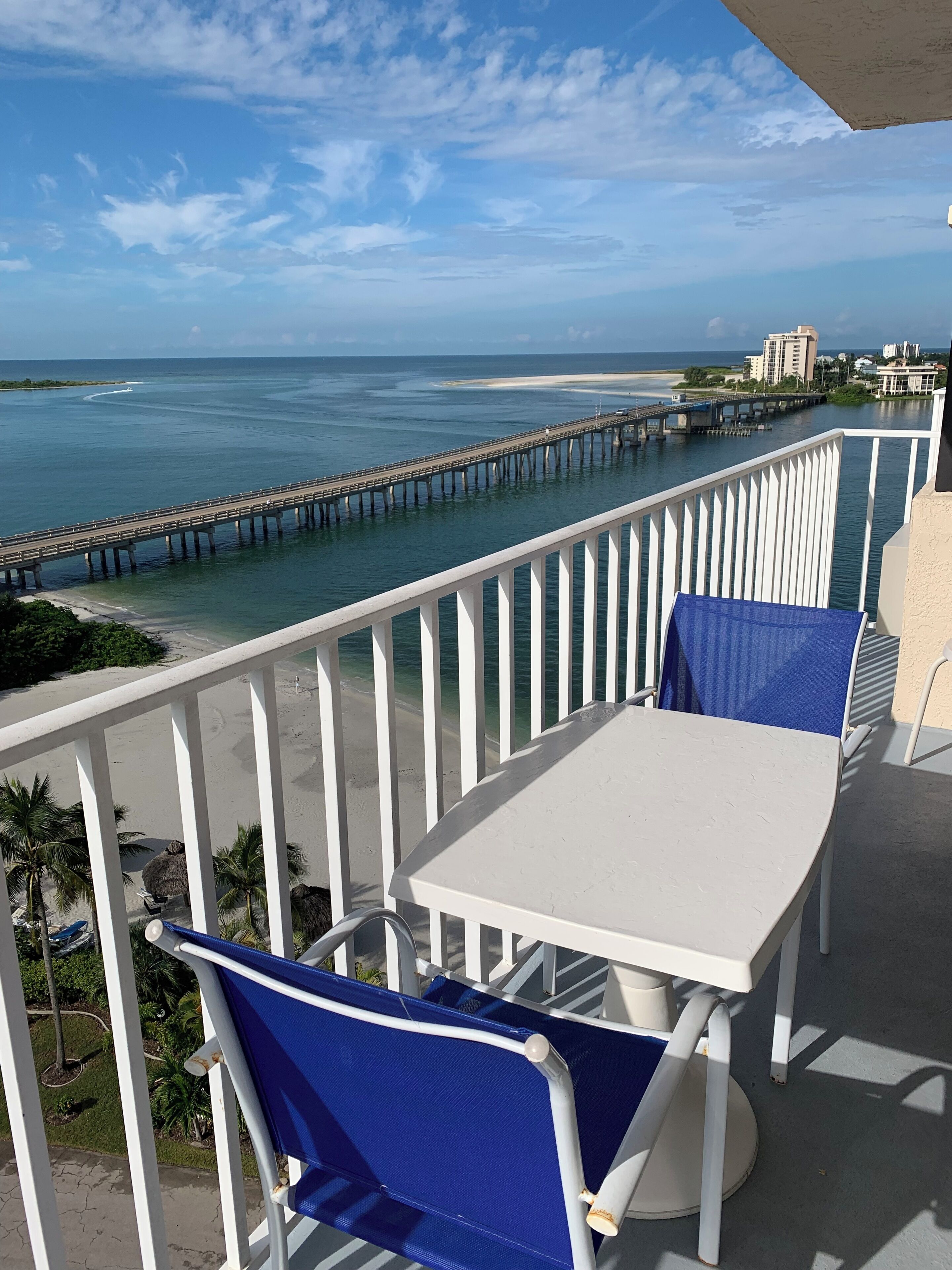 A balcony with a white table and blue chairs, overlooking a beach and a pier.