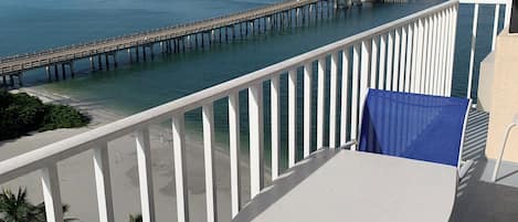 A balcony with a white table and blue chairs, overlooking a beach and a pier.