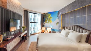 A modern hotel room with a large bed, a TV mounted on the wall, a desk with a chair, and a view of palm trees through the window. - Apec Mandala Chambay Mui Ne - Oceanic Studio (Mui Ne)