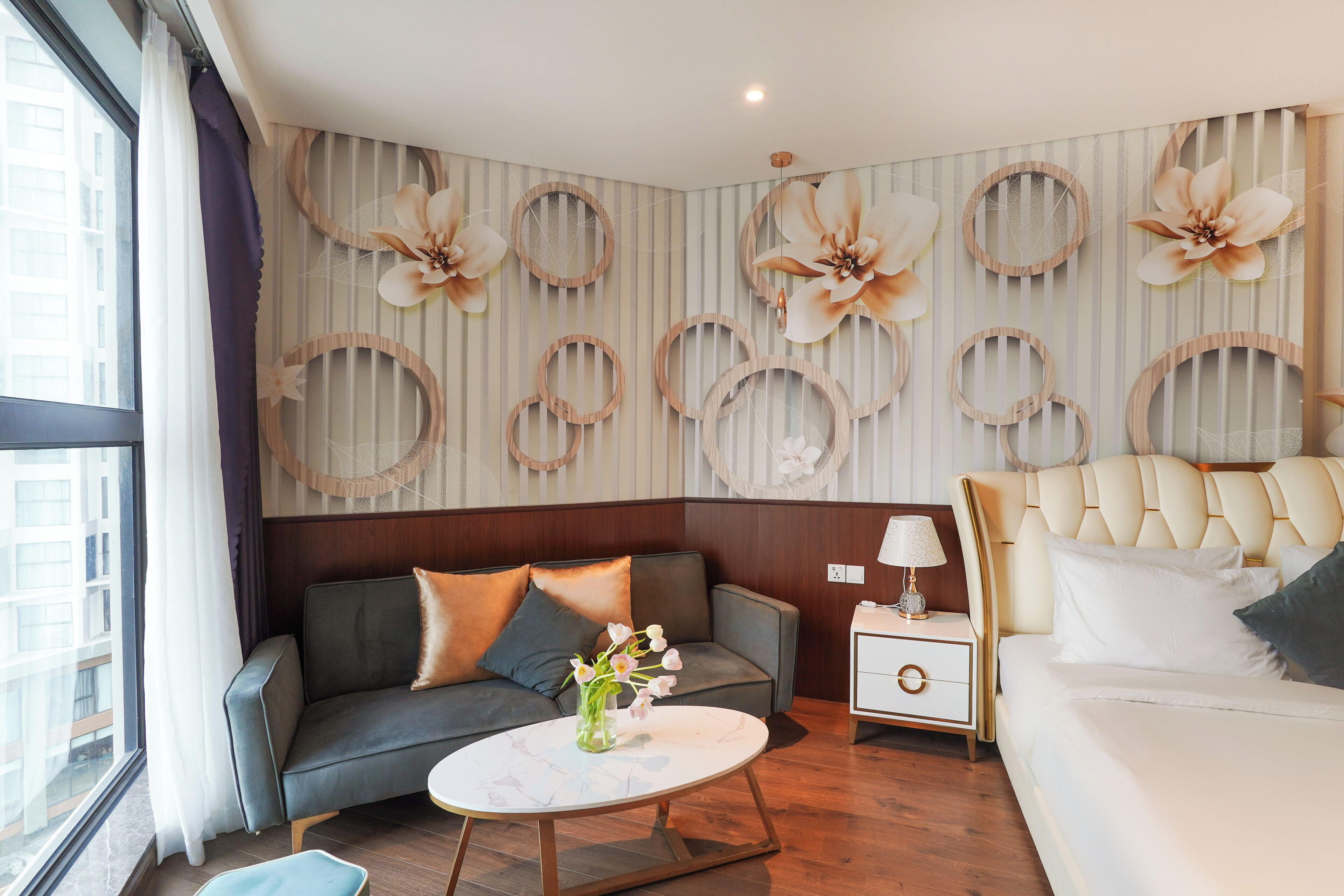 A modern hotel room with a sofa, a bed, a nightstand, and a wall decorated with circular frames and floral patterns.