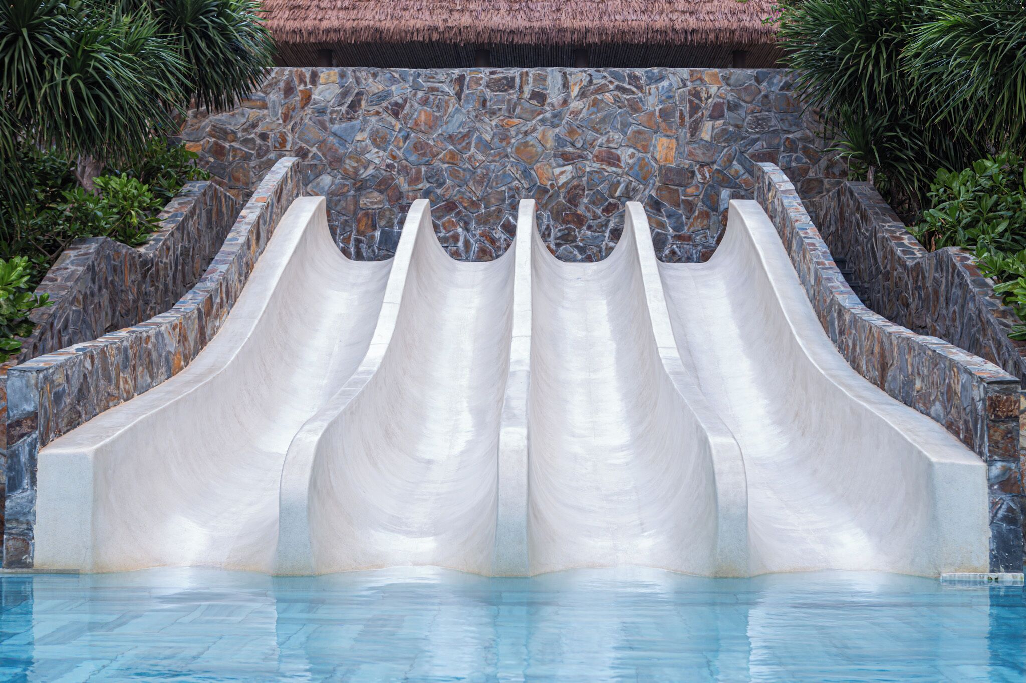 Une piscine dotée de quatre toboggans, entourée d’un mur de pierre et de plantes tropicales.