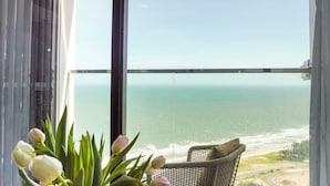 A balcony with a view of the ocean, a wicker chair with a cushion, and a vase of tulips. - Apec Mandala Chambay Mui Ne - Oceanic Studio (Mui Ne)