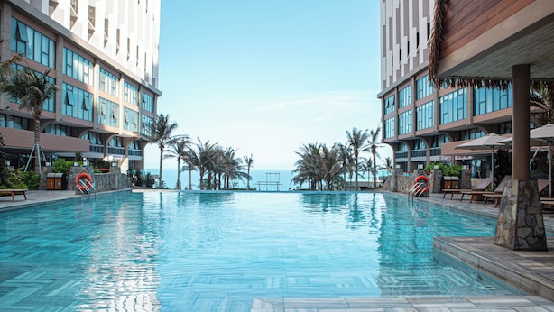 A swimming pool with a view of the ocean, surrounded by palm trees and modern buildings. - Apec Mandala Chambay Mui Ne - Oceanic Studio (Mui Ne)