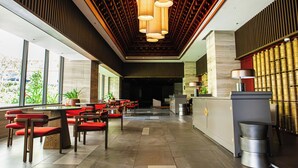 A modern hotel lobby with a long reception desk, multiple seating areas with red chairs, and a large window offering a view of greenery outside. - Apec Mandala Chambay Mui Ne - Oceanic Studio (Mui Ne)