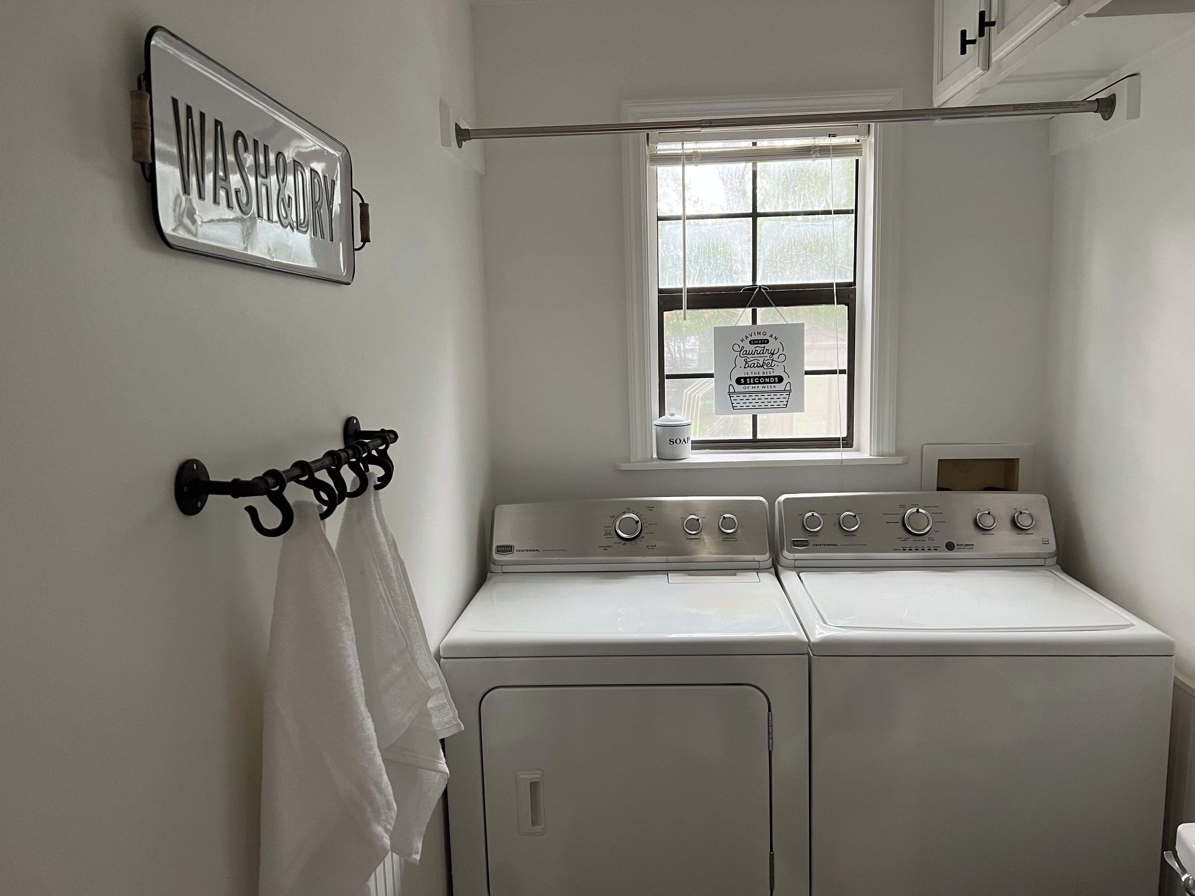 Een wasserette met twee voorladers en drogers, een raam met uitzicht op bomen, en een 'WASH & DRY' bord aan de muur.