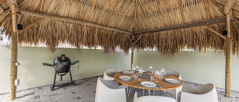 An outdoor dining area with a thatched roof, a grill, and white plastic chairs.