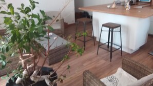 A modern living room with a wooden floor, a large potted plant, a wicker sofa, and a small bar area with stools and a countertop.