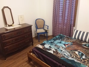 Un dormitorio con un aparador de madera, una silla azul, una cama con colcha estampada y un cojín a rayas.