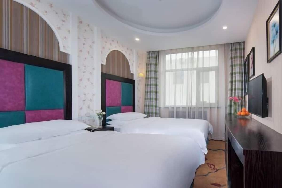 A hotel room with two beds, a desk, a TV, and a large window with curtains.