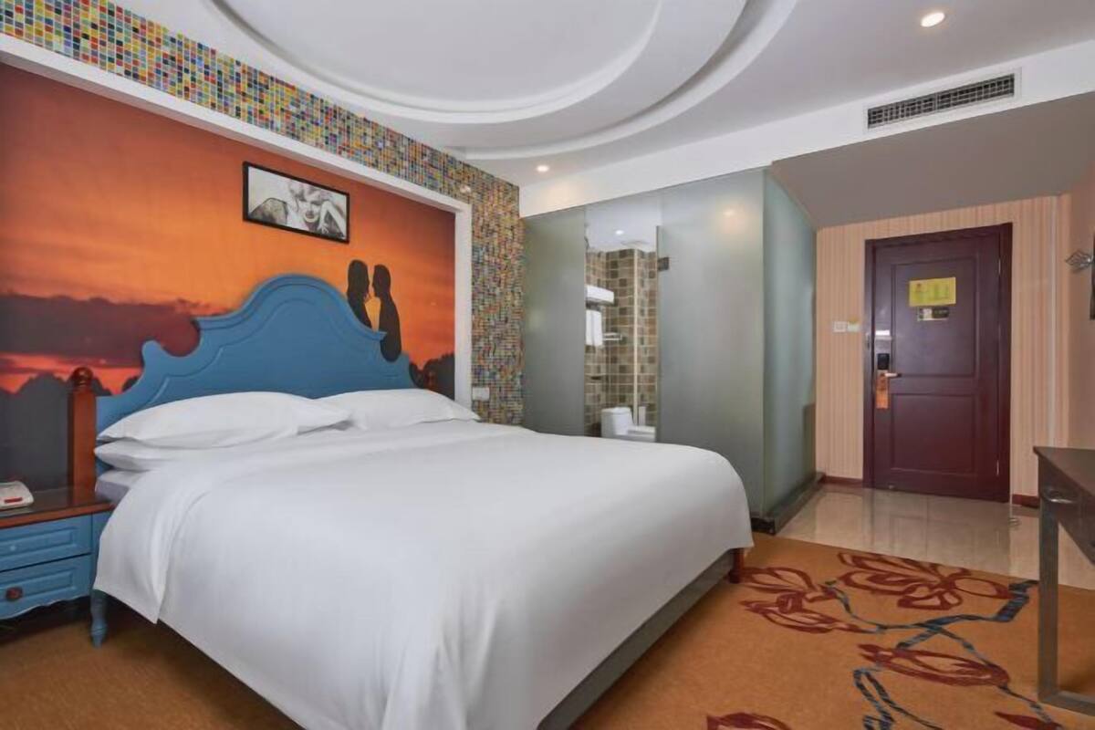 A hotel room with a bed, a nightstand, a bathroom with a shower, and a door.