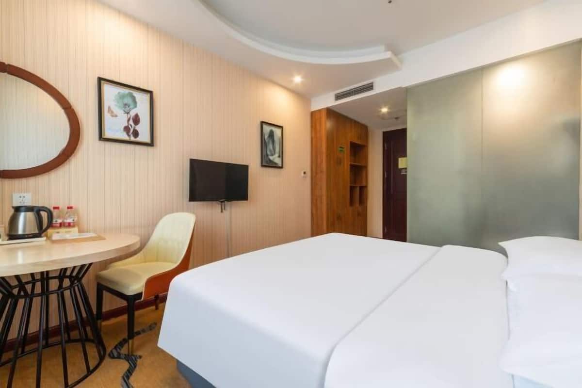 A hotel room with a bed, a TV, a small table with a kettle, and a chair.