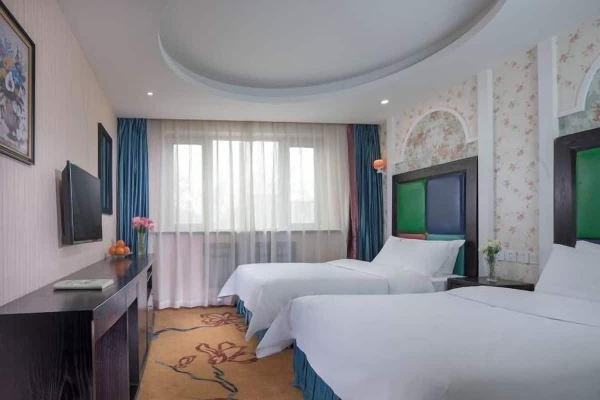 A hotel room with two beds, a desk with a flat-screen TV, a large window with sheer curtains, and a floral wallpaper design.