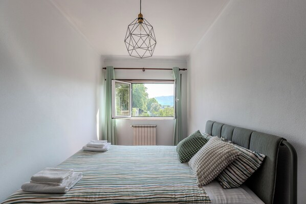 Una cama bien hecha con ropa de cama a rayas, una lámpara colgante de diseño geométrico y vistas a la vegetación a través de una ventana con cortinas.