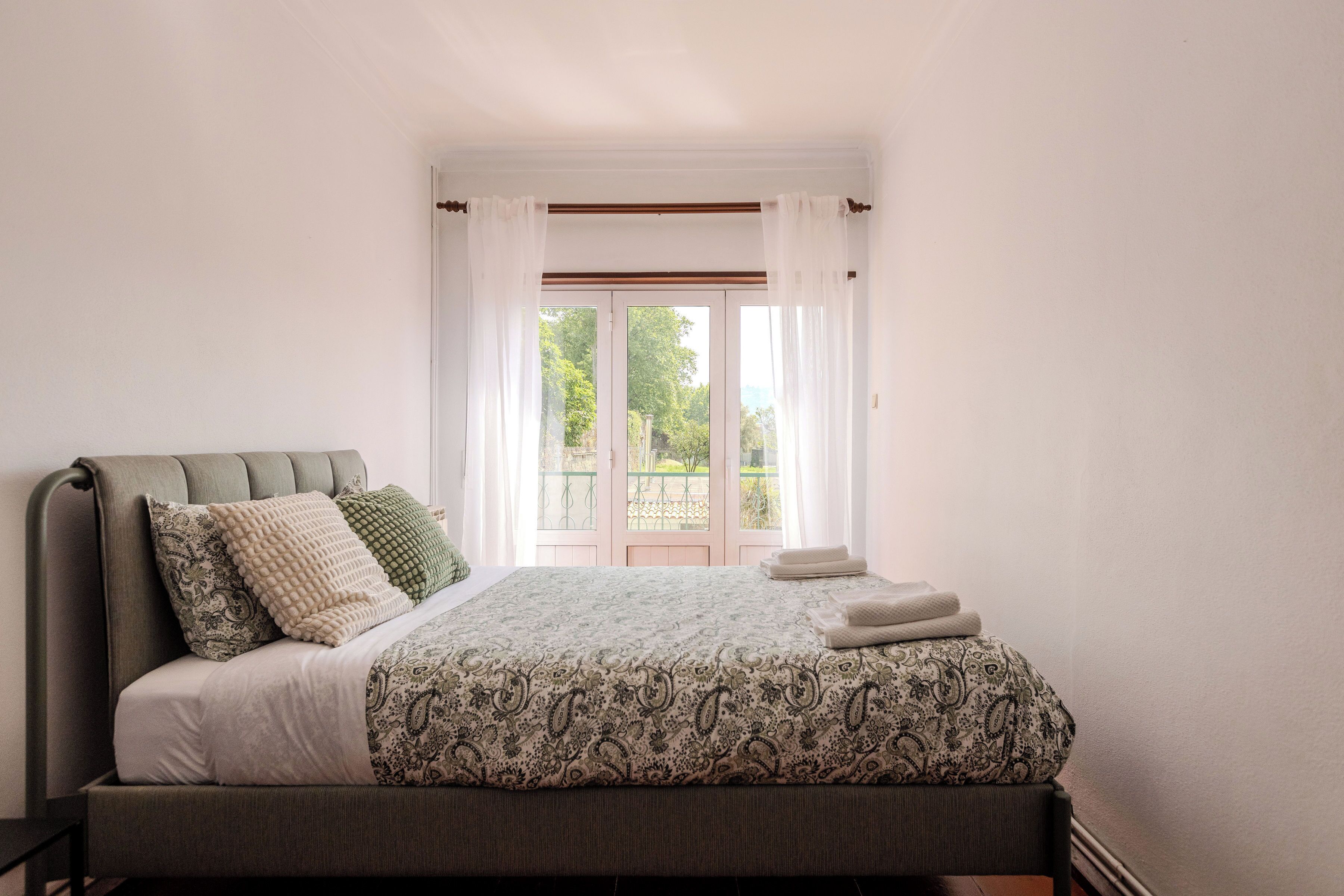 A neatly made bed with a patterned bedspread, a bedside table, and a view of a garden through a window with curtains.