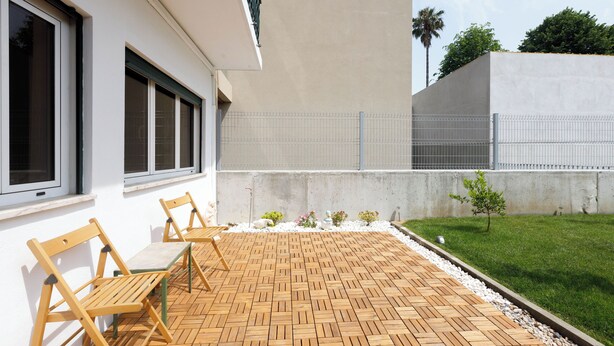 Un patio con suelo de madera, mobiliario exterior y una pequeña zona ajardinada con plantas y una palmera.