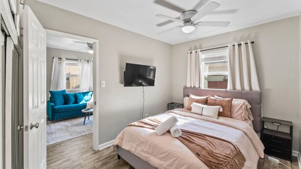 Un dormitorio con cama, televisor, ventilador de techo y vistas a un salón con sofá azul.